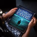 Indigo Code Hardback - Woman on chair holds a book
